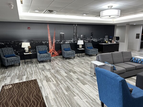 A modern hotel lobby with a cityscape view, grey patterned armchairs, a grey sofa, and a bar area. - Garner Hotel Oklahoma City - Quail Springs by IHG (Oklahoma City)