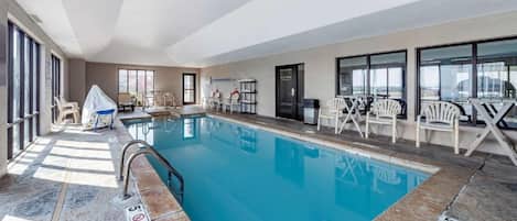 Una piscina interior con agua cristalina, rodeada de sillas y mesas blancas, y grandes ventanales en un lateral.