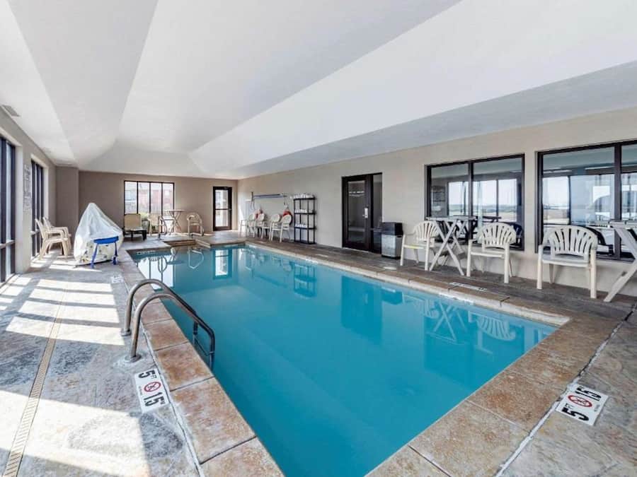 An indoor swimming pool with clear water, surrounded by white chairs and tables, and large windows on one side.
