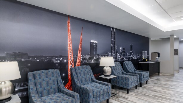 Un hall d’hôtel orné d’une fresque représentant un paysage urbain, avec quatre fauteuils à motifs, un appareil à café et une lampe posée sur une table d’appoint.