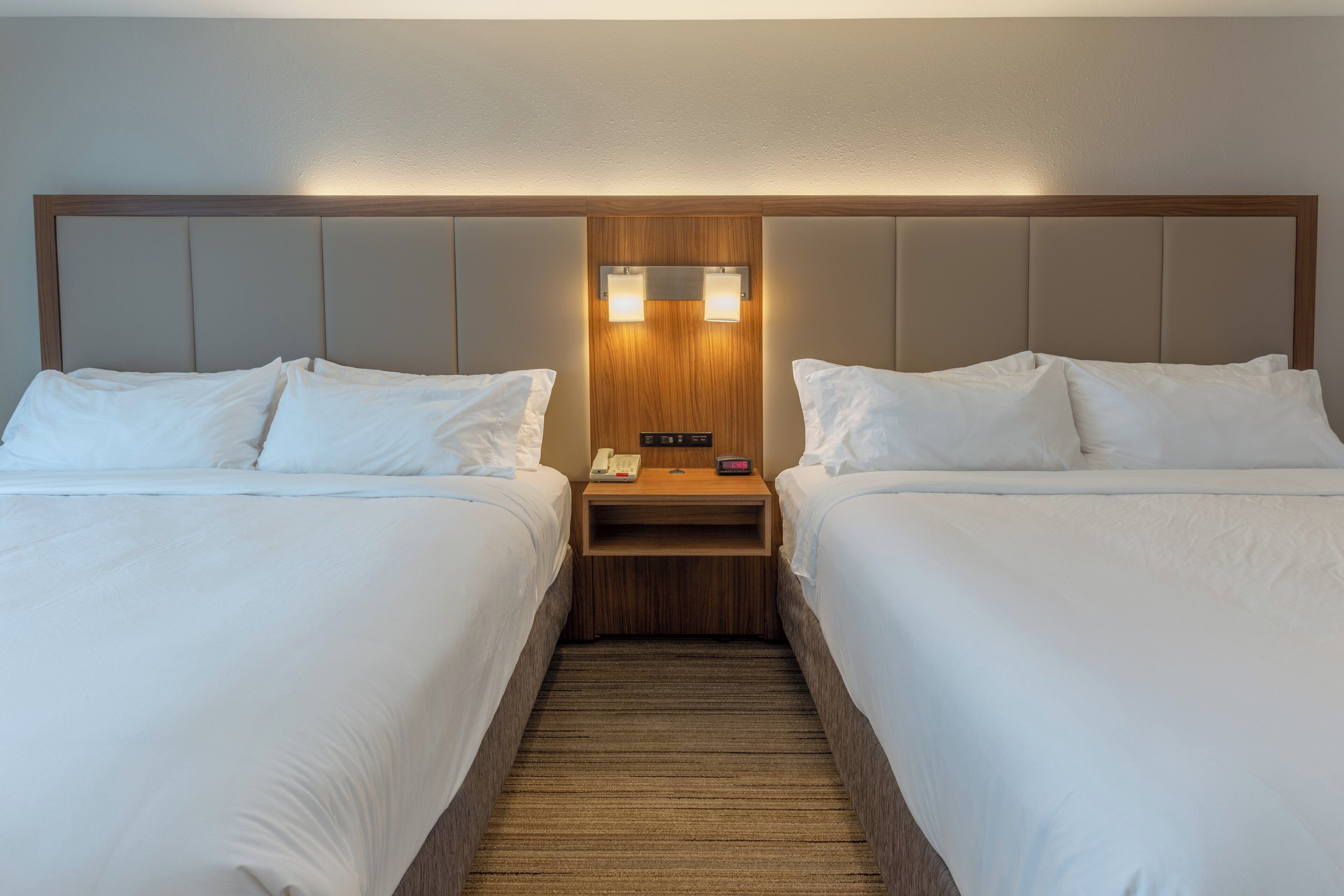 Habitación de hotel con dos camas, cabecero de madera, mesita de noche con teléfono y reloj digital, y piso alfombrado.