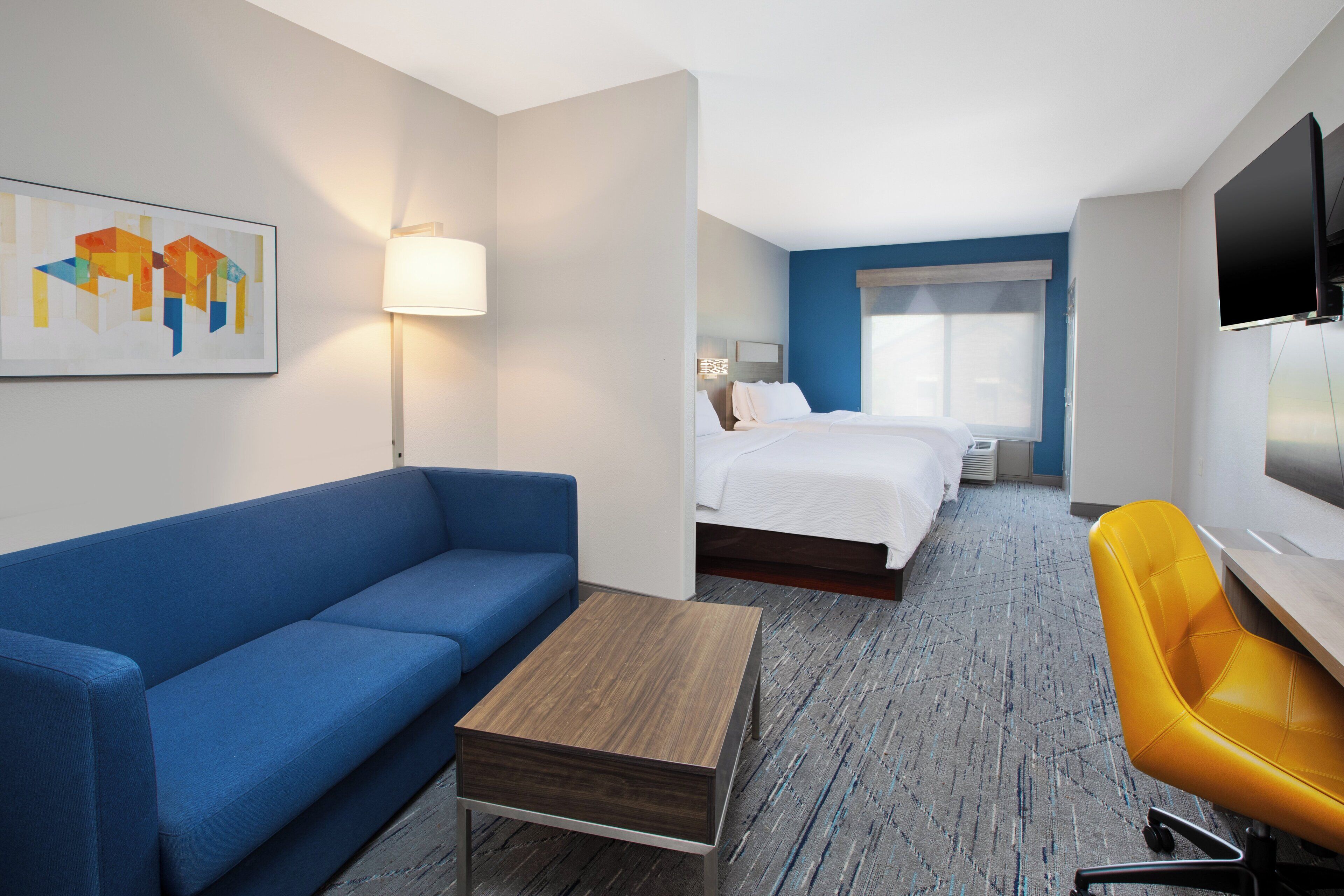 A hotel room with a blue sofa, a wooden coffee table, a yellow chair, a bed with white bedding, a TV mounted on the wall, and a painting on the wall.