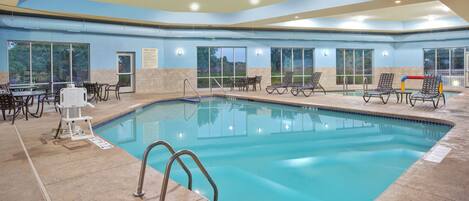 An indoor swimming pool with clear blue water, surrounded by lounge chairs and tables.