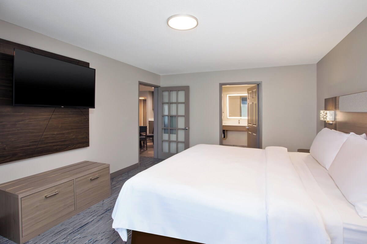Een moderne hotelkamer met een groot bed, een flatscreen televisie aan de muur, een houten dressoir en een badkamer met spiegel en wastafel.