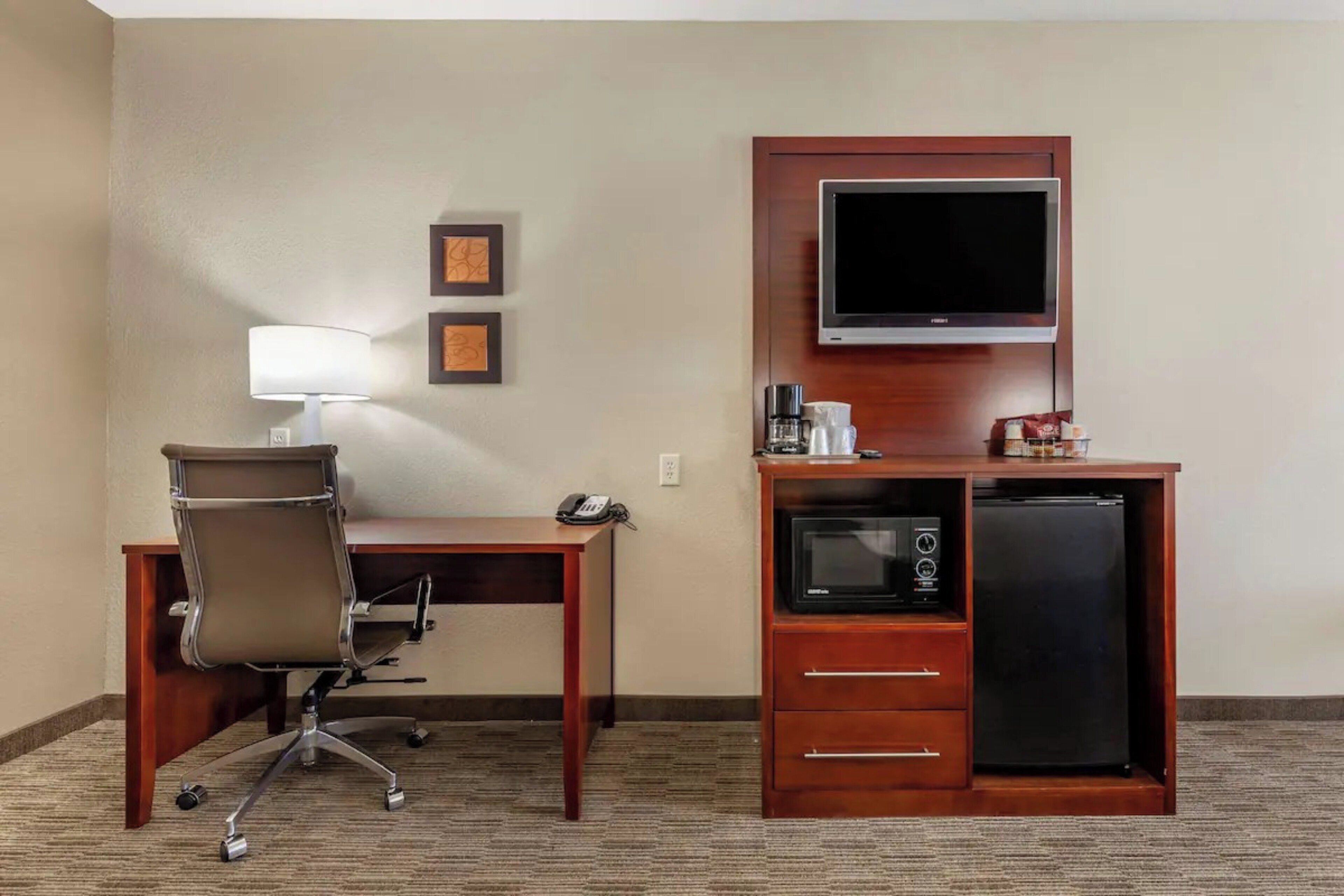 A hotel room with a desk, chair, lamp, TV, microwave, and refrigerator.