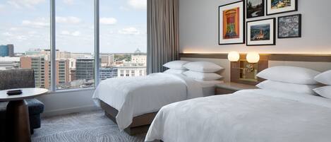A hotel room with two beds, a desk, and a large window offering a city view.
