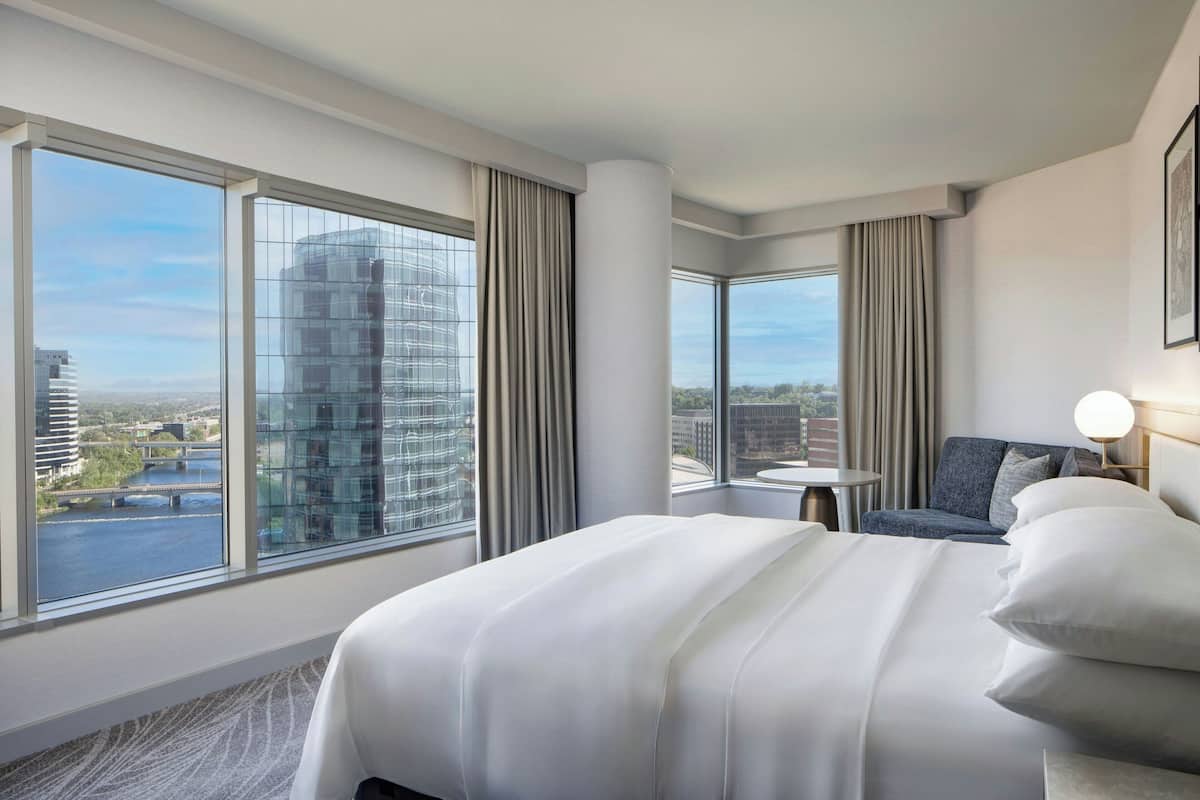 Camera d'albergo con un letto grande, una poltrona, un tavolino e vista sulla città attraverso ampie finestre.
