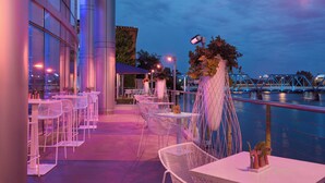 Un espace extérieur aménagé pour les repas, avec des chaises et des tables blanches, une plante décorative et une vue sur un pont et l’eau.
