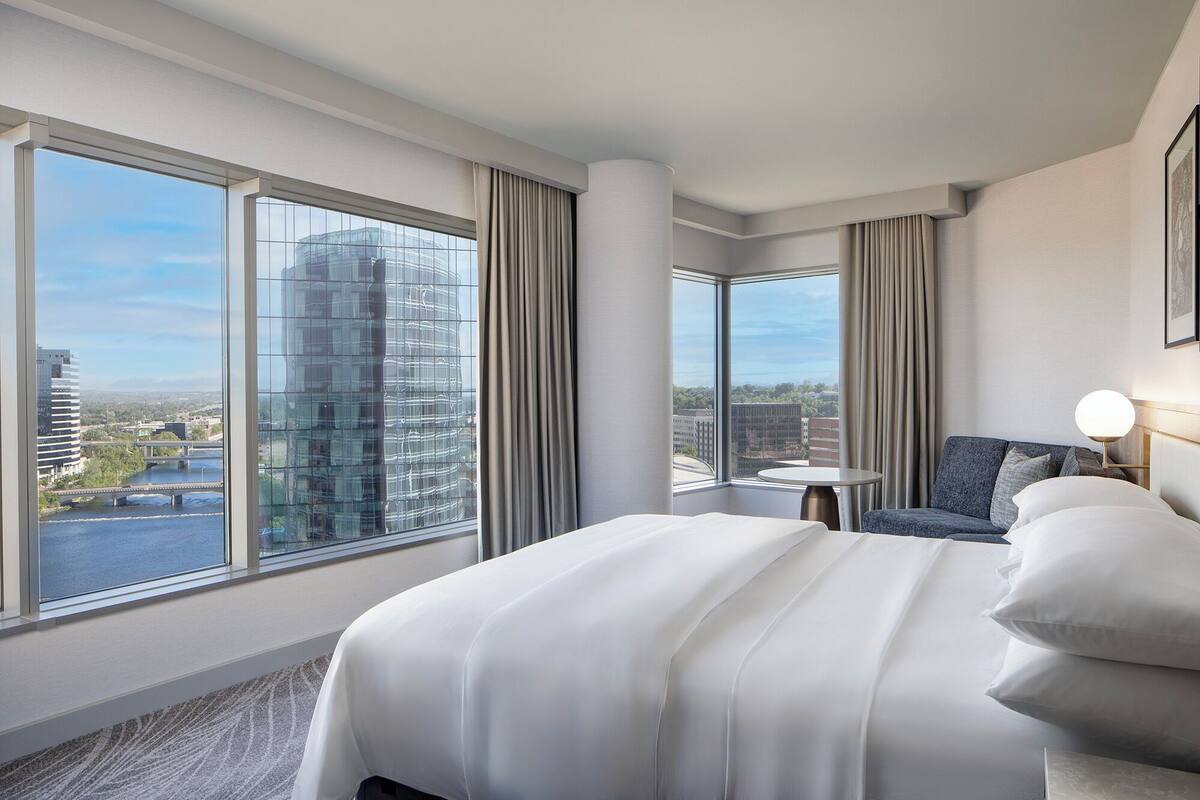 Una moderna camera d'albergo con un letto grande, un'area salotto e vista sulla città attraverso ampie finestre.