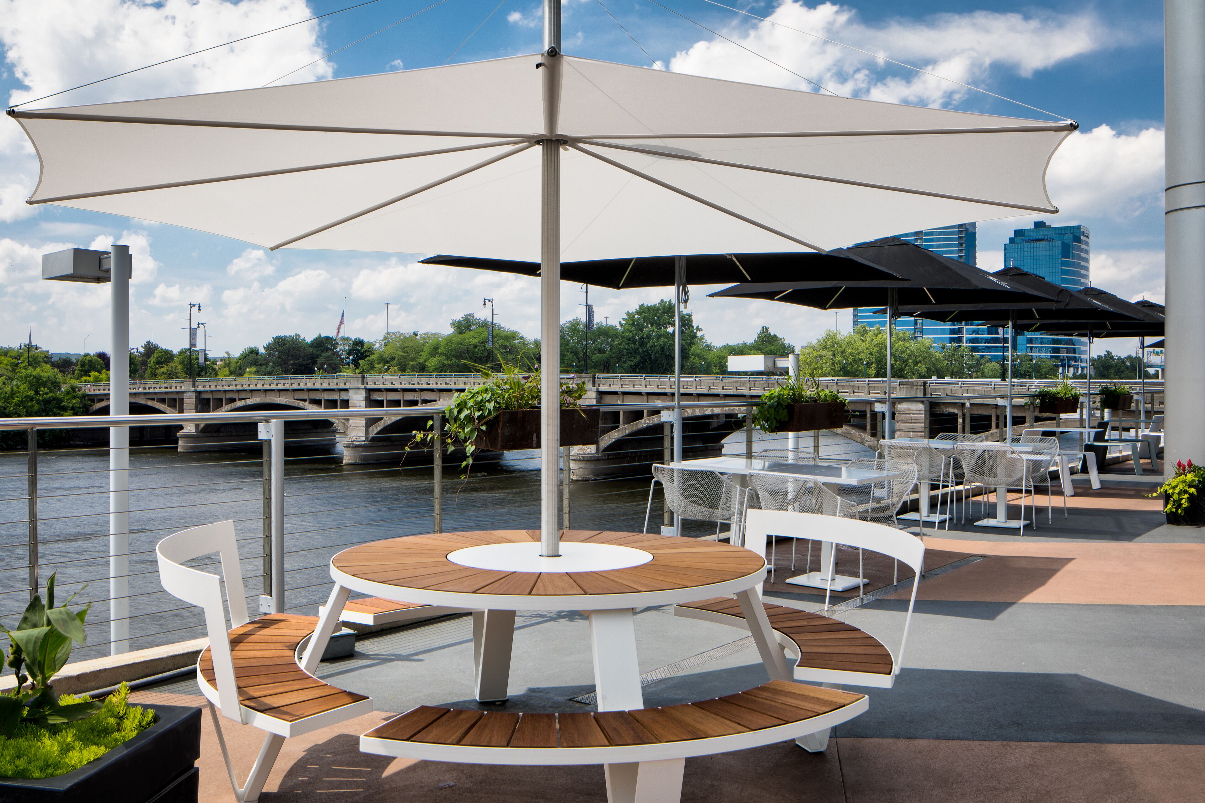 Een terras aan de rivier met een ronde houten tafel en witte stoelen, omringd door parasols en met uitzicht op een brug en gebouwen op de achtergrond.
