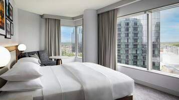 A modern hotel room with a large bed, a small table, and a view of the city through large windows.