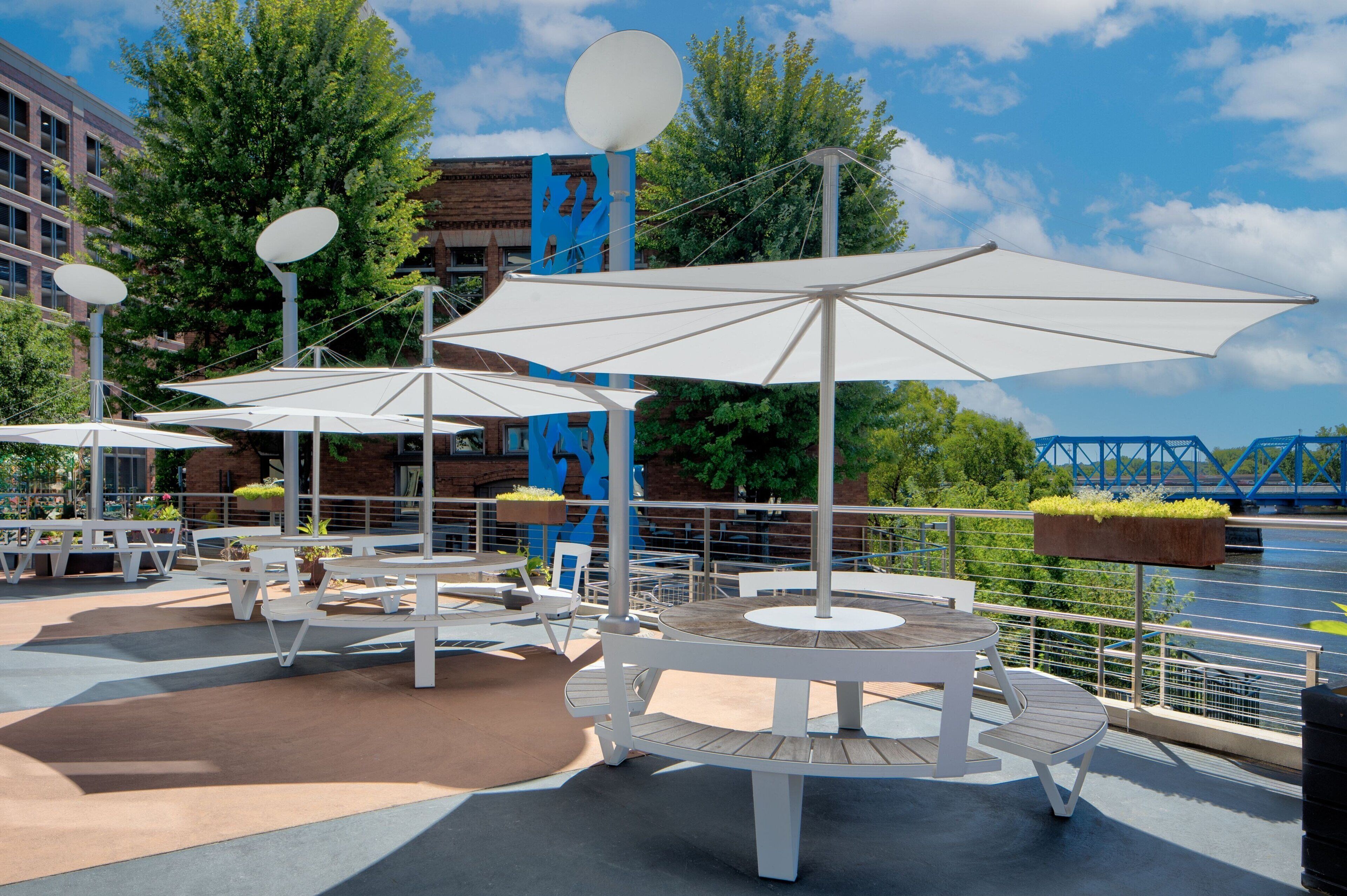 An outdoor patio area with white tables and umbrellas, surrounded by greenery and a view of a river.