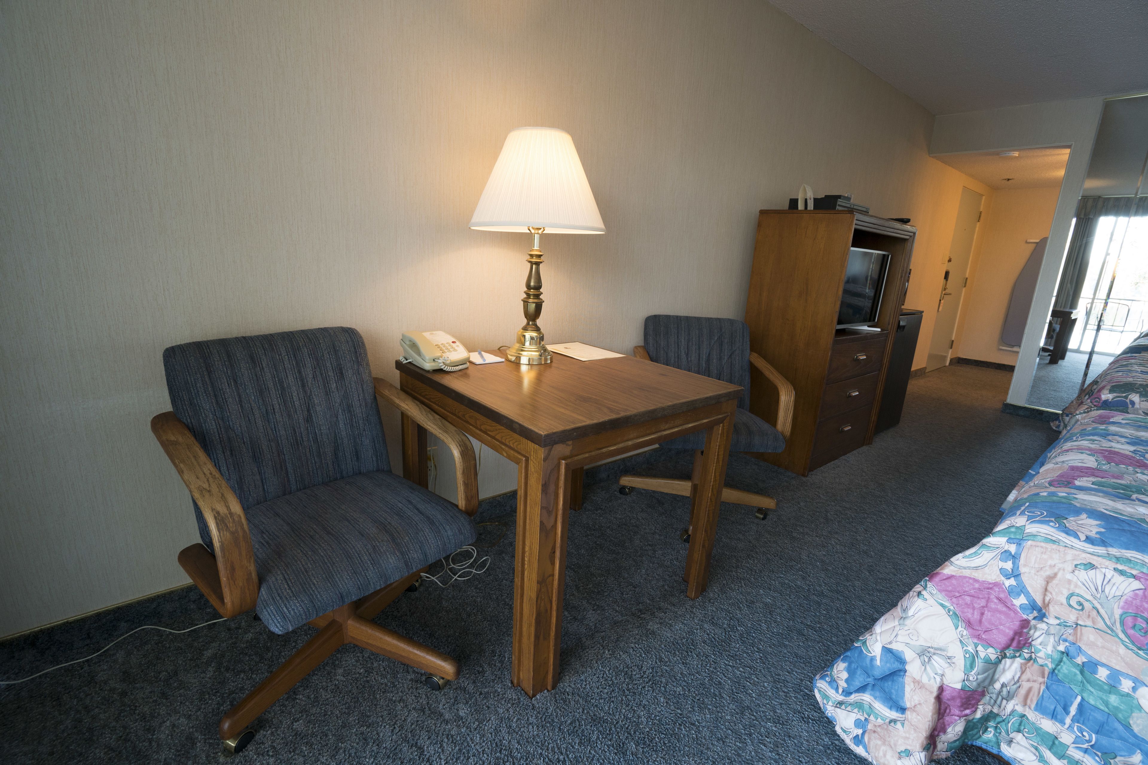 A hotel room with a wooden desk, two chairs, a lamp, a telephone, a television, and a bed with a colorful bedspread.
