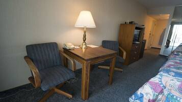 A hotel room with a wooden desk, two chairs, a lamp, a telephone, a television, and a bed with a colorful bedspread.