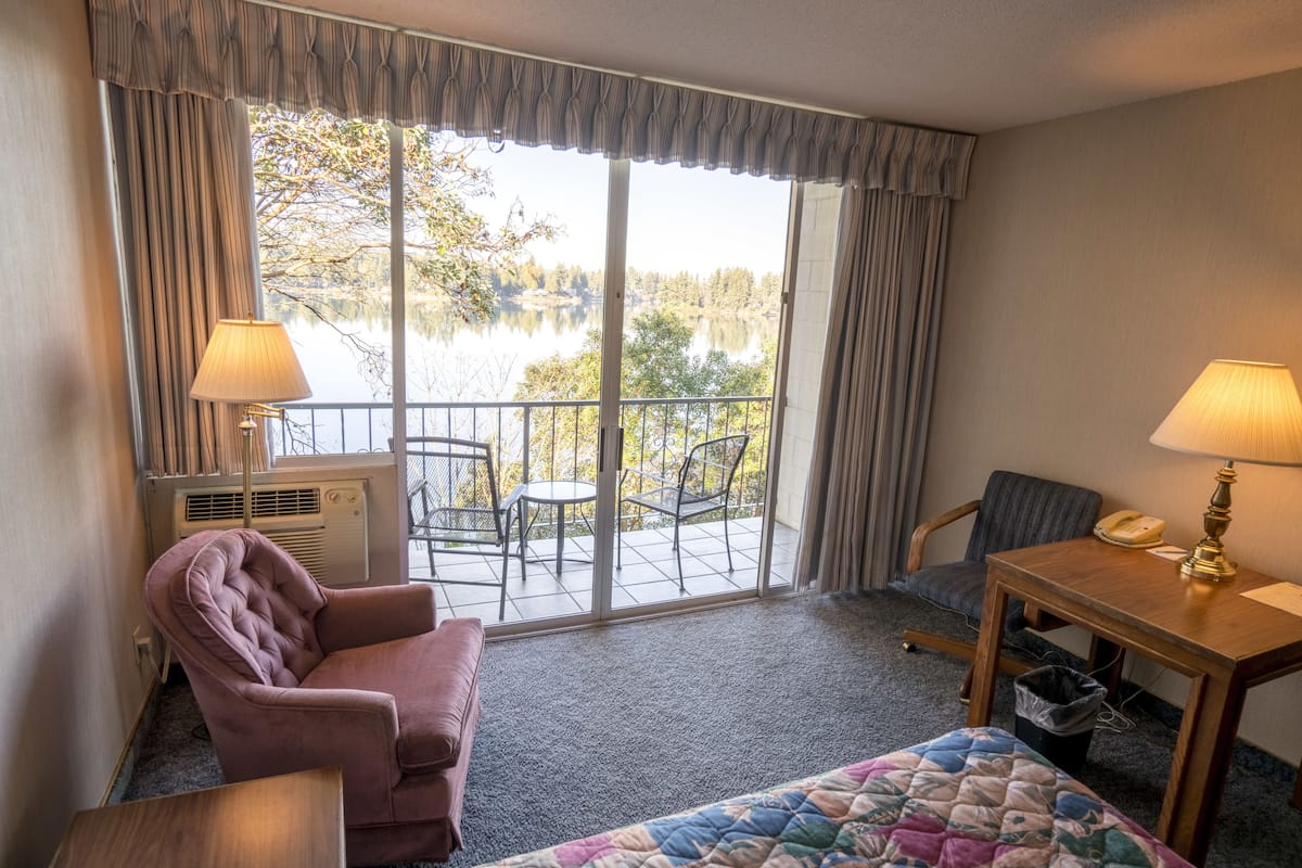 A hotel room with a bed, a chair, a table, a lamp, a phone, and a view of a lake through large windows.