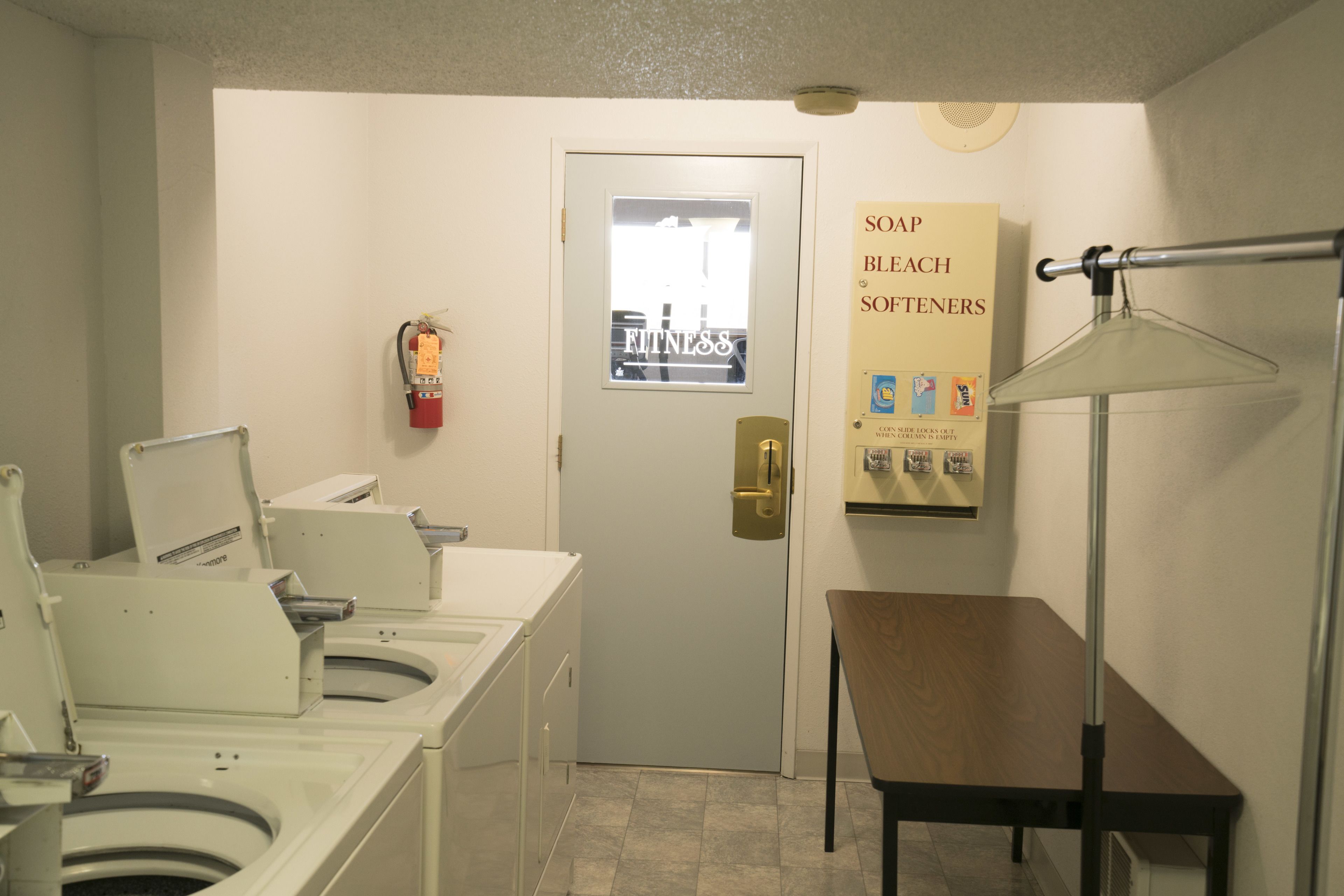 A laundry area with multiple washing machines, a door with a poster, and a table with a lamp.