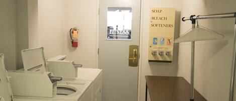 A laundry area with multiple washing machines, a door with a poster, and a table with a lamp.