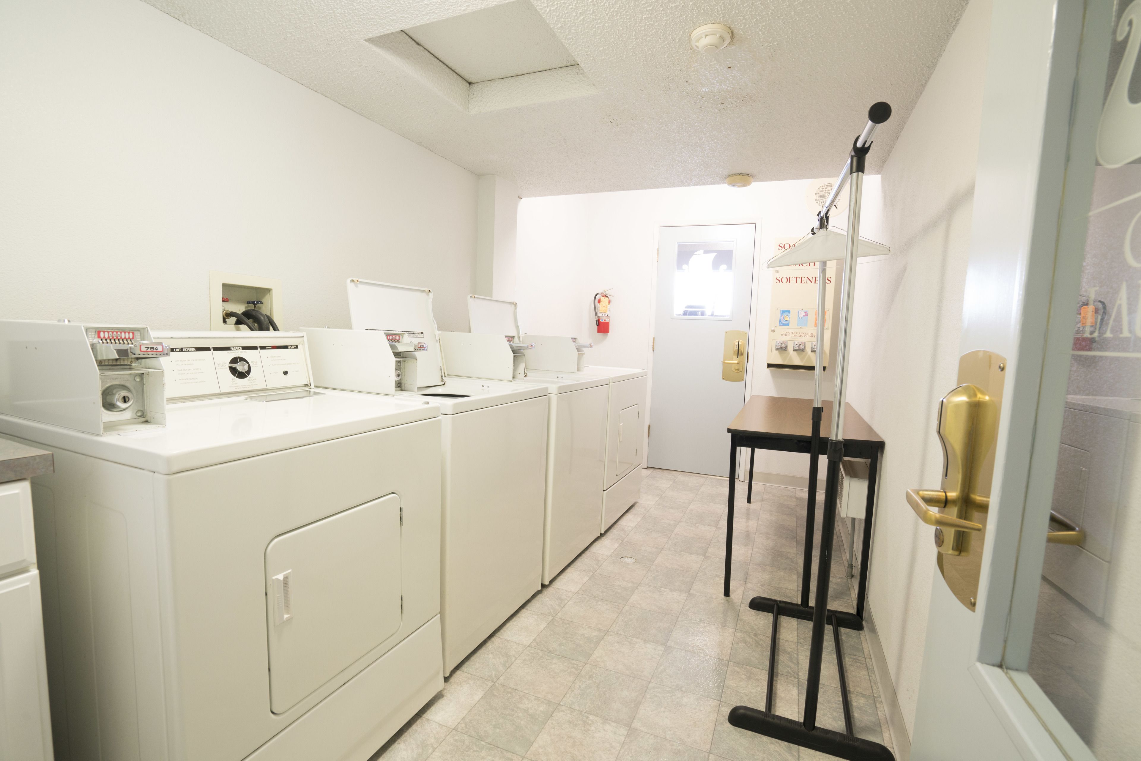 A laundry room with white appliances, a small table with a lamp, and a door with a golden handle.
