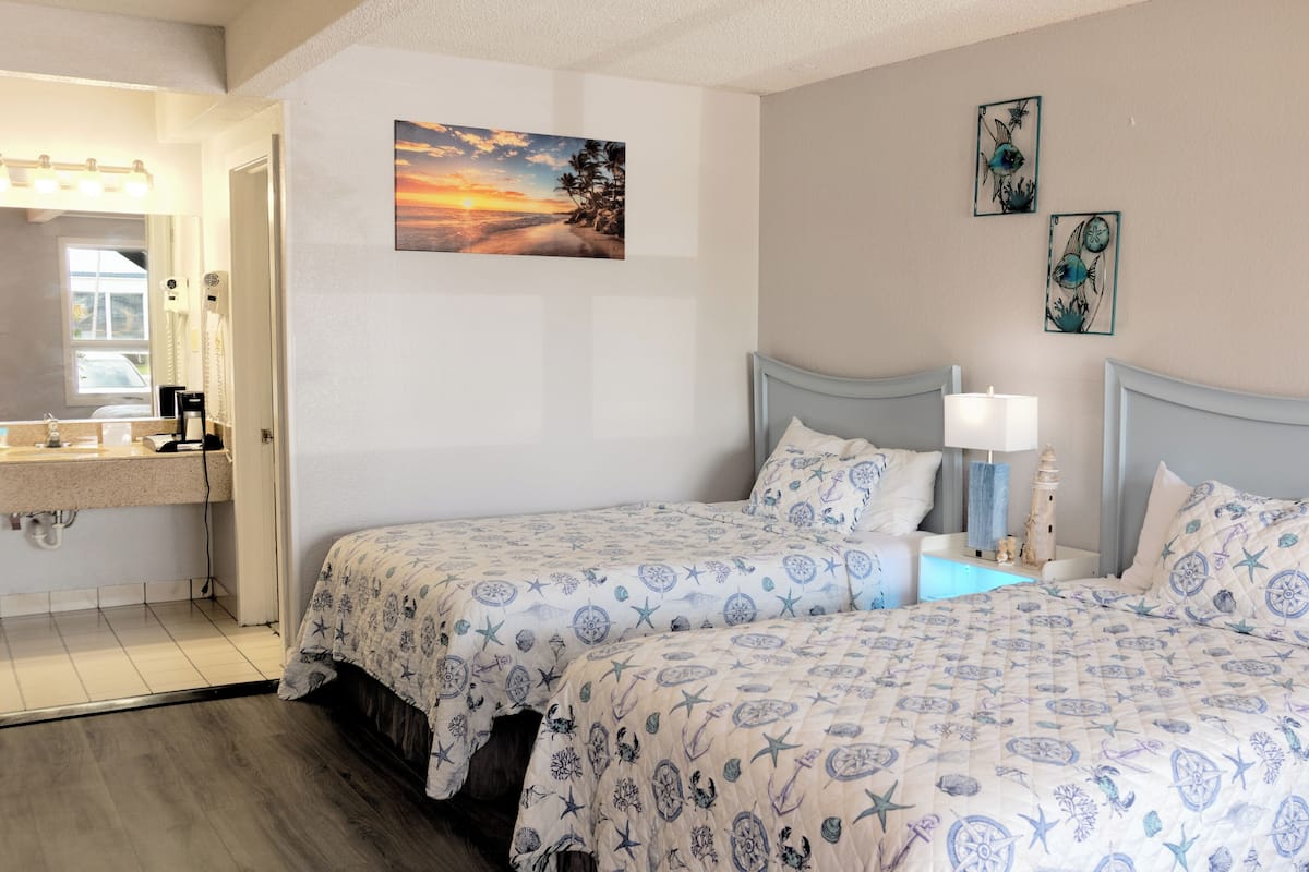 A hotel room with two beds, a bathroom with a sink, and a painting of a sunset.