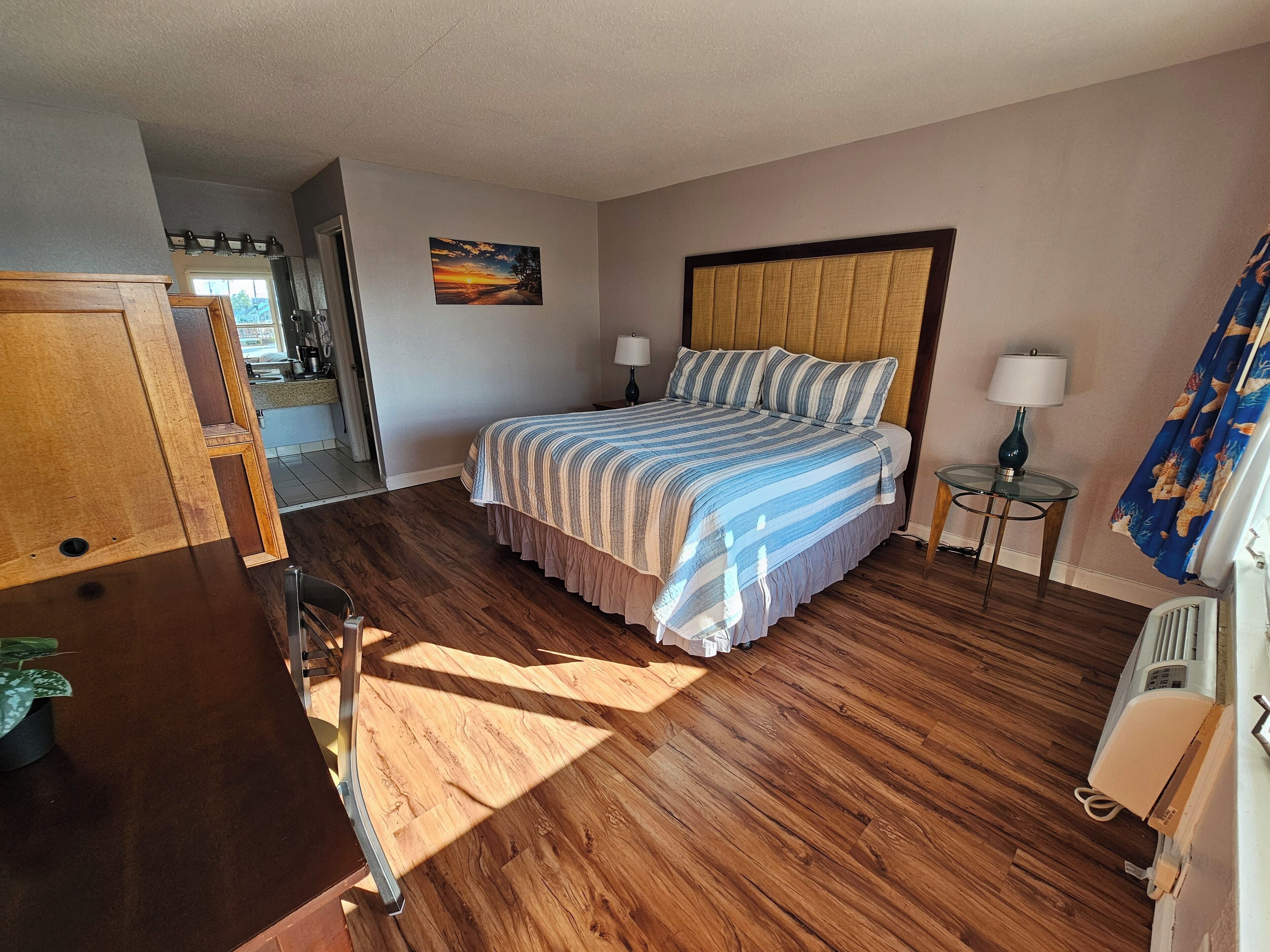 Une chambre avec un plancher en bois, un lit avec une literie à rayures, une table de chevet avec une lampe et un tableau accroché au mur.