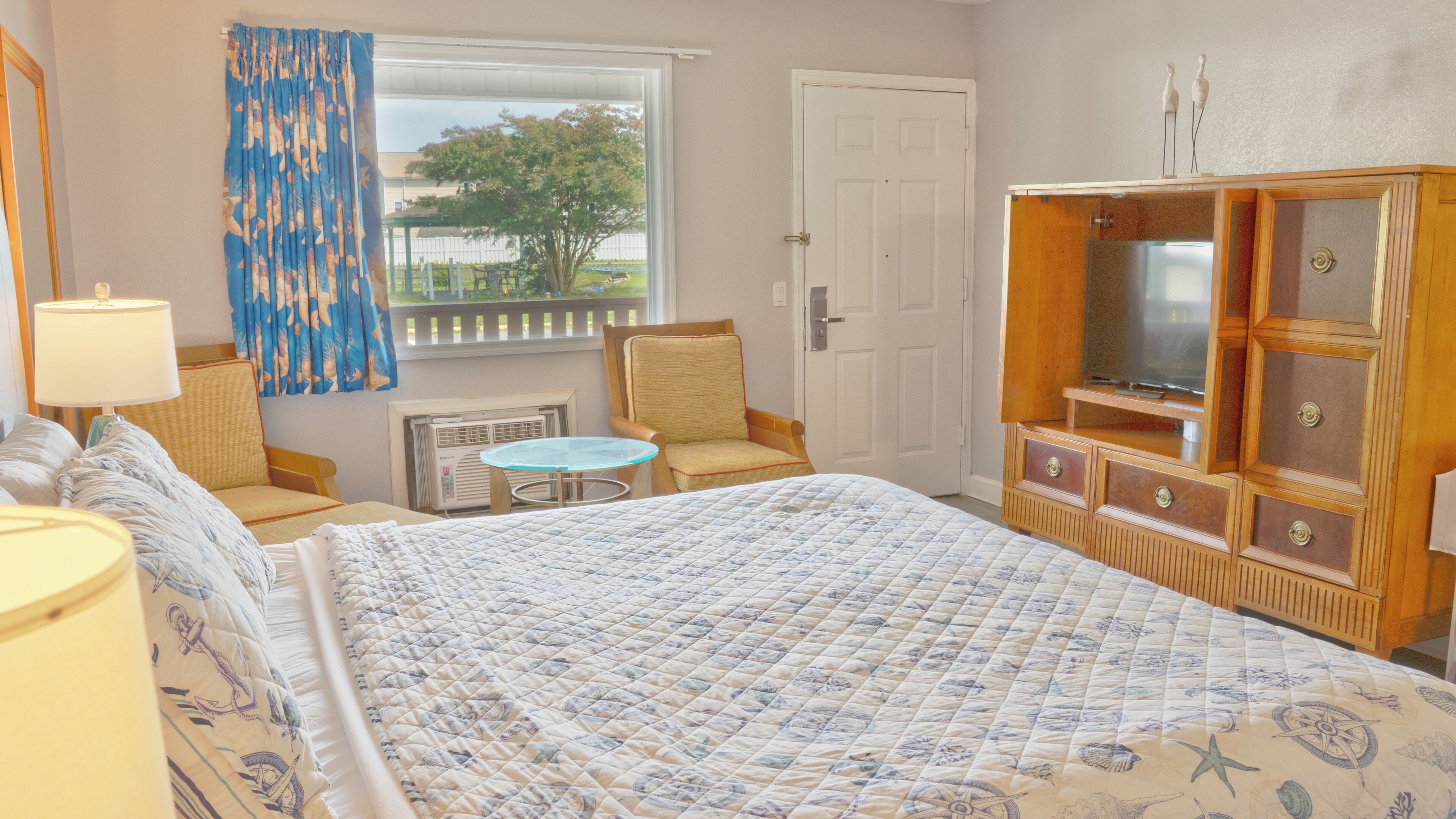 A hotel room with a bed, a chair, a small table, a TV cabinet, and a window with curtains.