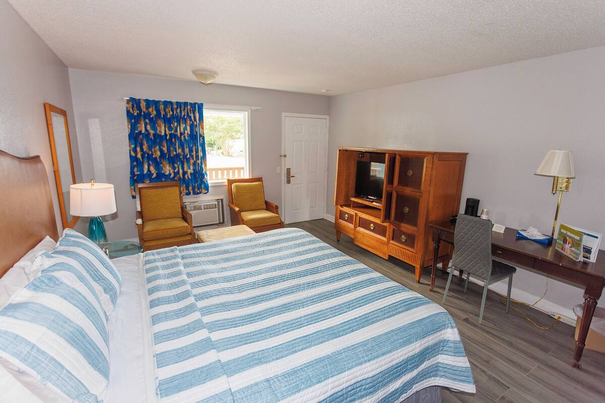 A hotel room with a bed, two chairs, a desk with a computer, a TV, and a window with curtains.