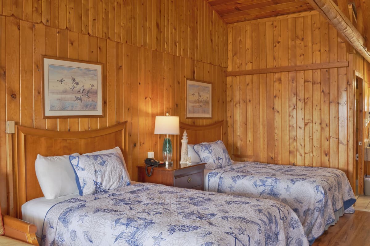 A wooden cabin room with two beds, a nightstand, a lamp, and framed artwork on the walls.