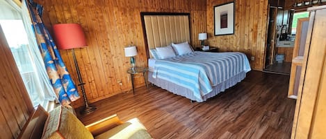 Een houten kamer met een bed, een stoel, een tafel met een lamp, en een keuken die zichtbaar is door een open deur.