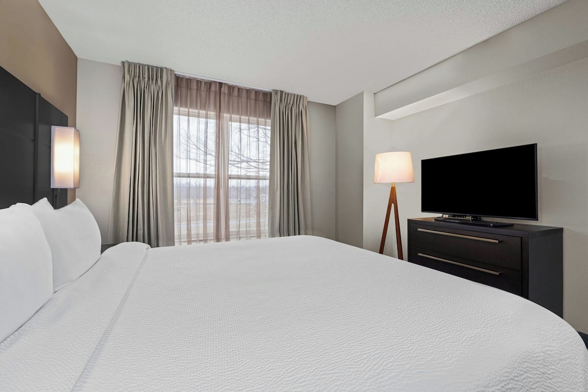 A hotel room with a large bed, a flat-screen TV, a bedside table with a lamp, and a window with curtains.