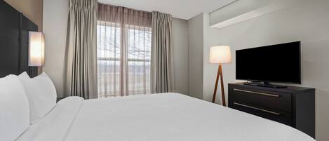 A hotel room with a large bed, a flat-screen TV, a bedside table with a lamp, and a window with curtains.