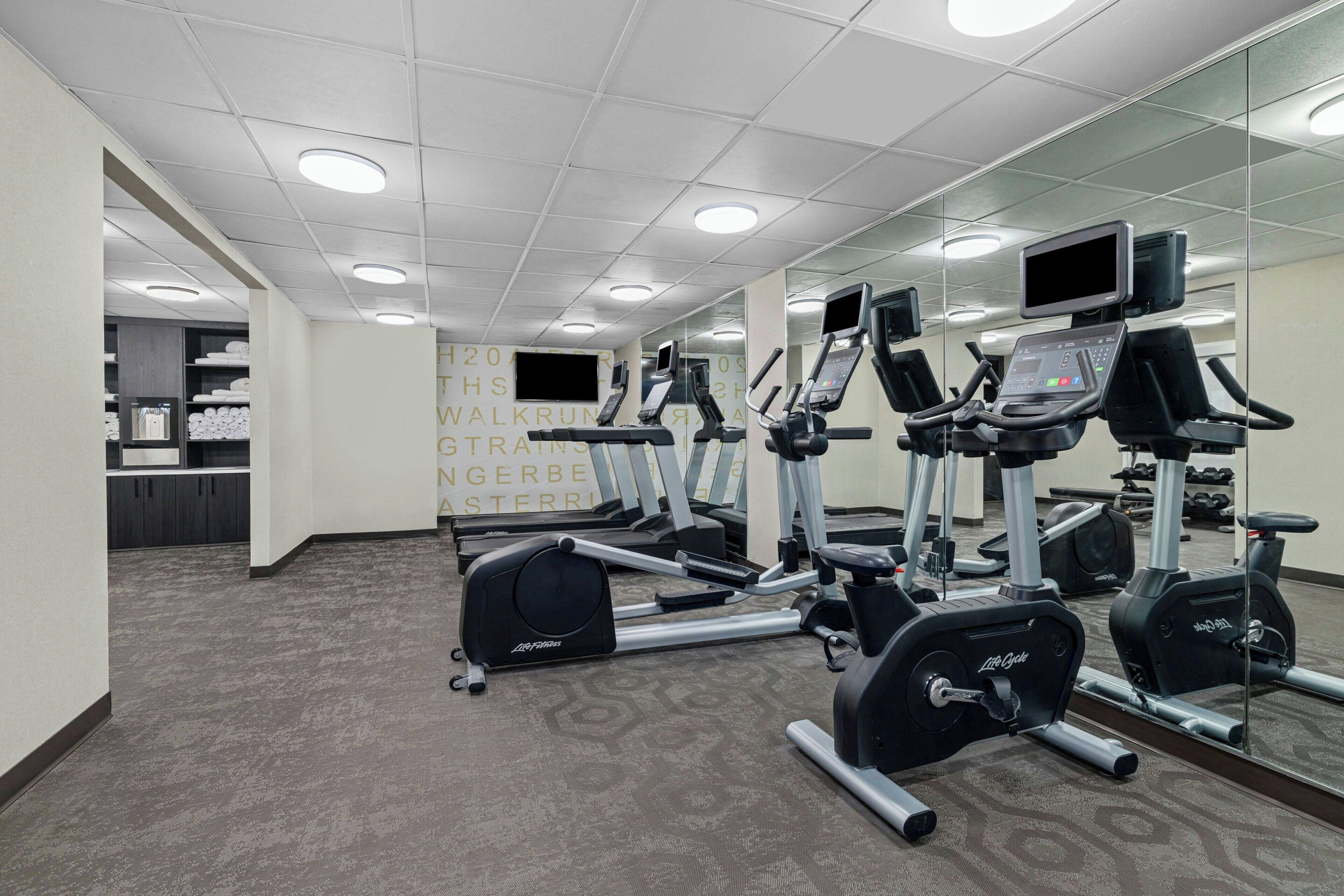 Ein Fitnessstudio mit Laufbändern, Crosstrainern und Gewichten, ausgestattet mit einer Spiegelwand und Teppichboden.