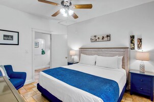 A hotel room with a large bed, a ceiling fan, a nightstand with a lamp, a blue chair, and artwork on the walls.