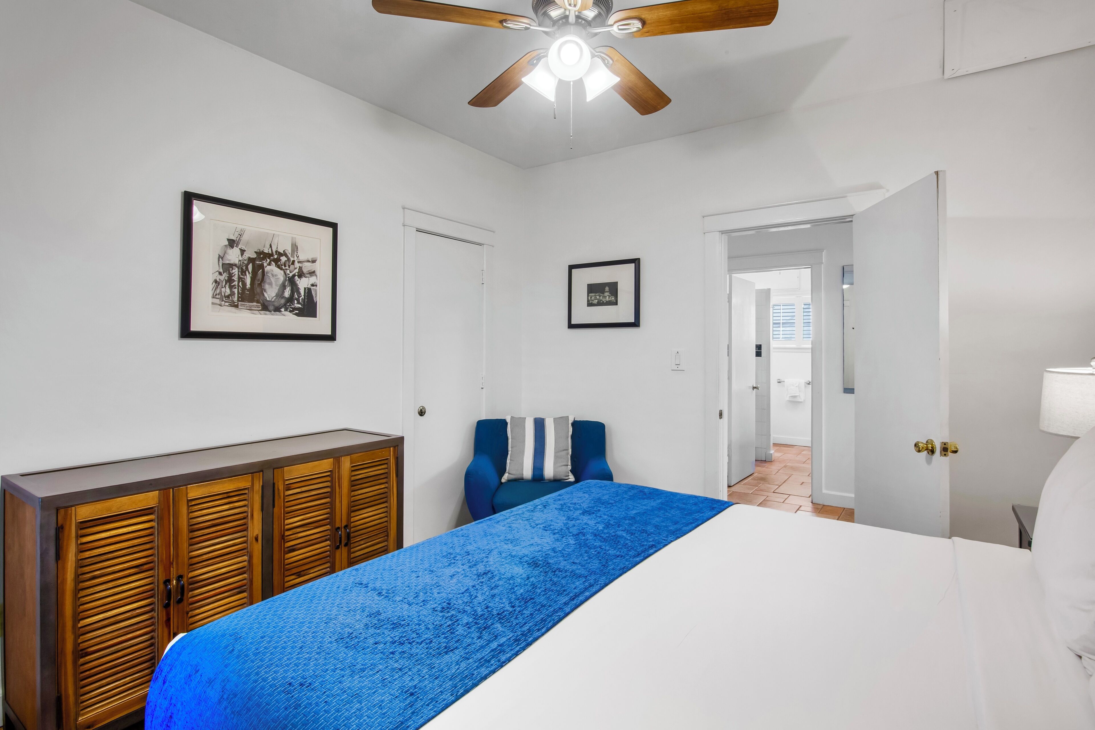 A bedroom with a bed, a wardrobe, two framed pictures, a ceiling fan, and a door leading to another room.