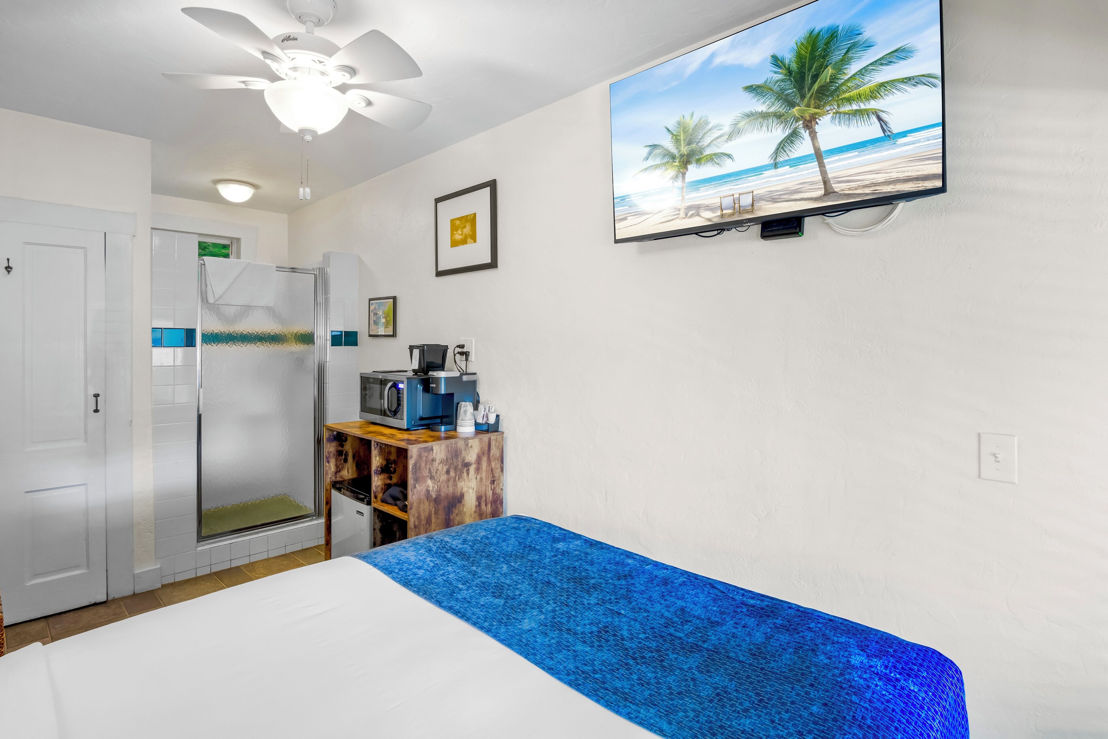 A hotel room with a bed, a ceiling fan, a TV displaying a beach scene, a small desk with a coffee maker, and a door leading to a shower area.