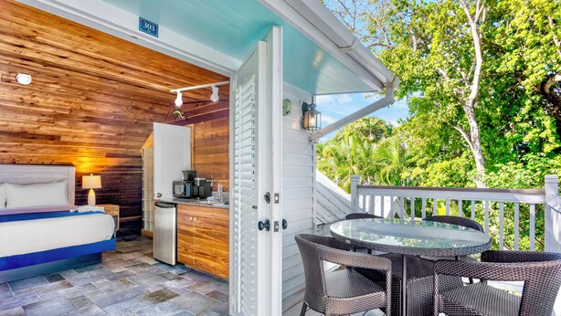 A bedroom with a bed, bedside table, lamp, and a small kitchenette. There is a balcony with wicker chairs and a glass table. - Eden House (Key West)