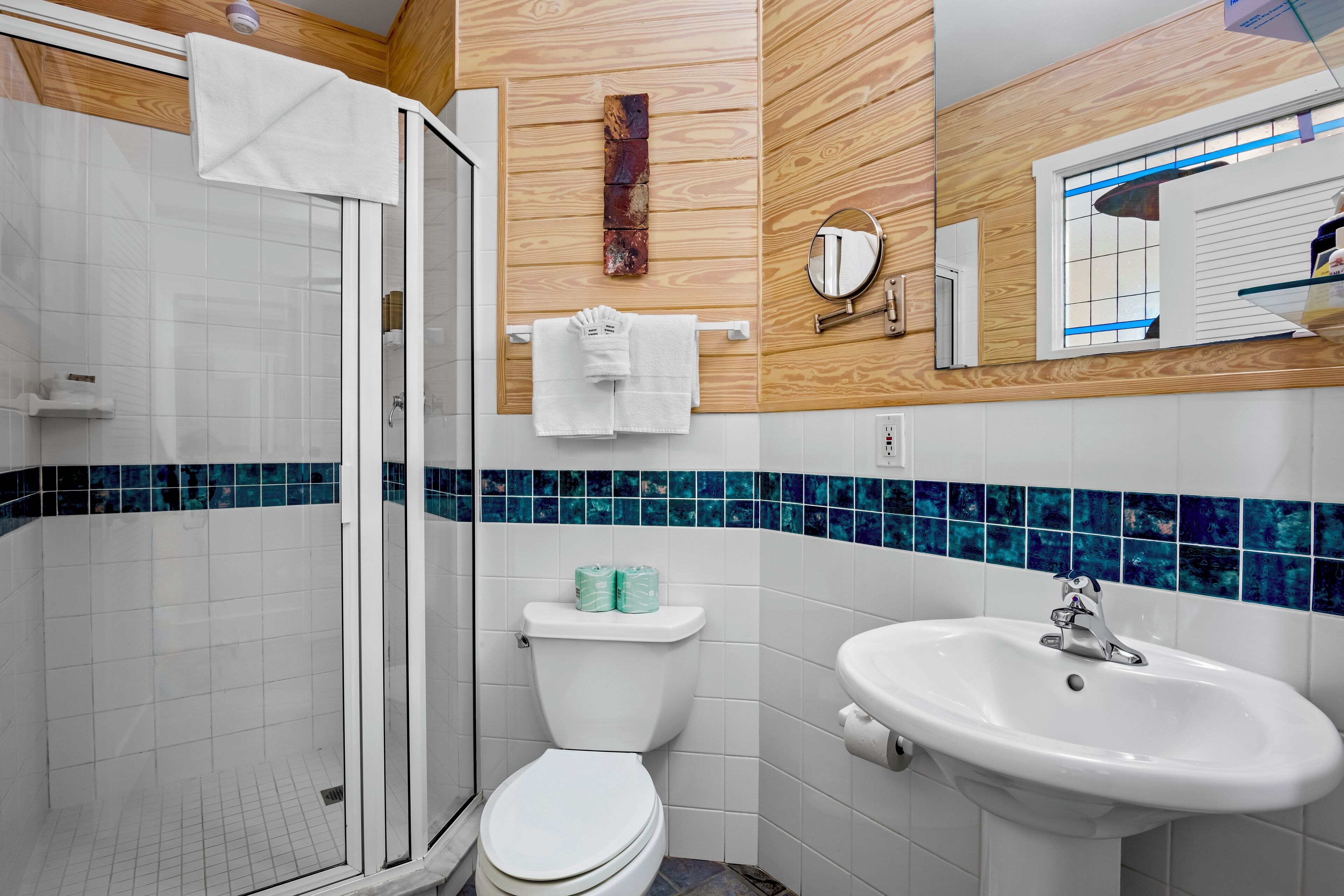 Un baño con cabina de ducha de vidrio, inodoro, lavamanos y pared de madera con detalles de azulejos azules.