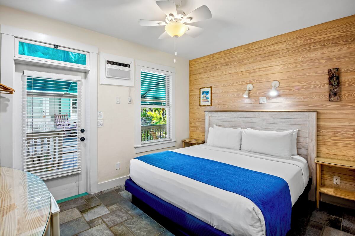A hotel room with a bed, a ceiling fan, a window with blinds, a wooden wall panel, and a small table.