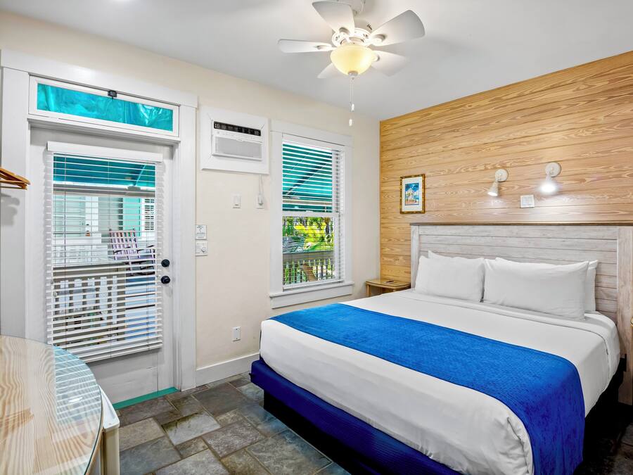 Ein Hotelzimmer mit Bett, Deckenventilator, Fenster mit Jalousien, einer Holzpaneelausstattung und einem kleinen Tisch.