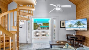 A spacious living area with a wooden staircase, a ceiling fan, a TV, and a view of the beach through an open door. - Eden House (Key West)