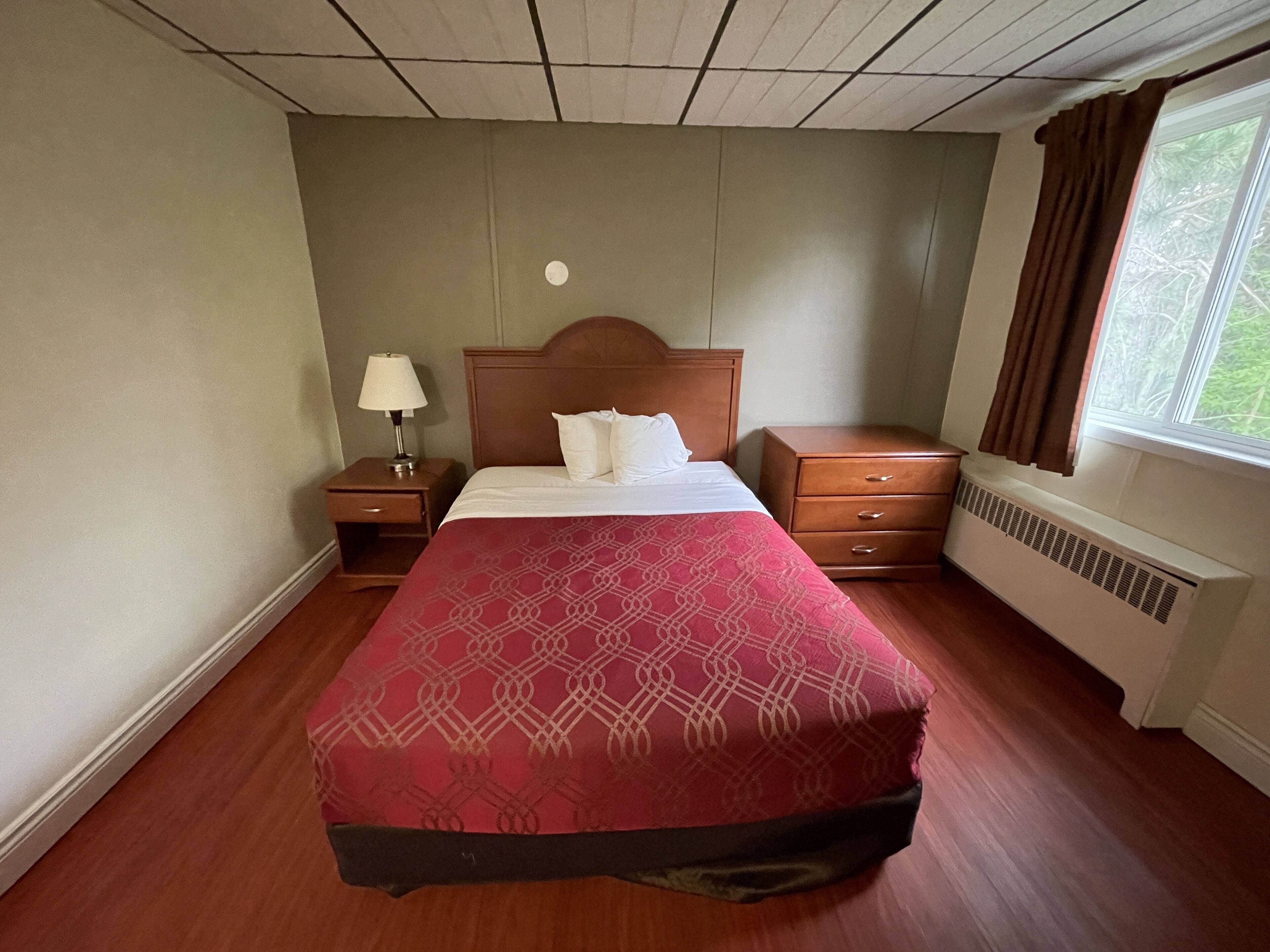 A hotel room with a large bed, a nightstand with a lamp, a dresser, and a window with curtains.