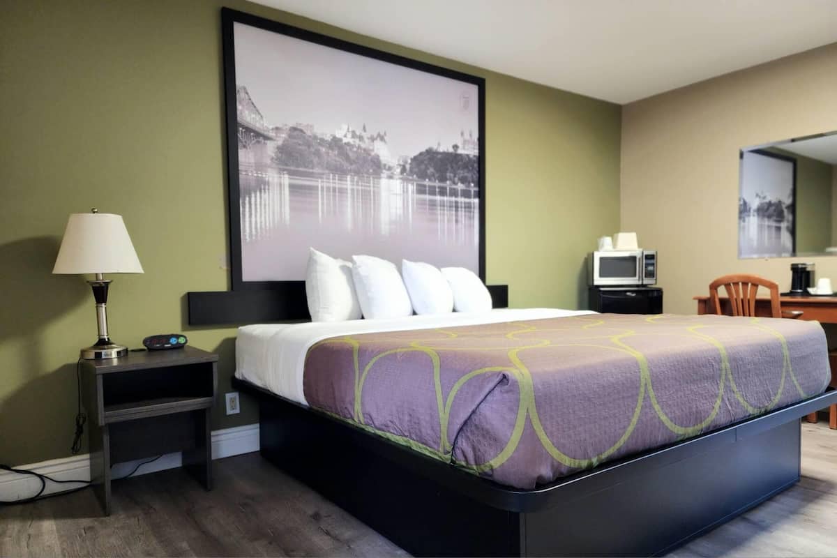A hotel room with a bed, a nightstand with a lamp, a desk with a computer, and a large framed picture on the wall.