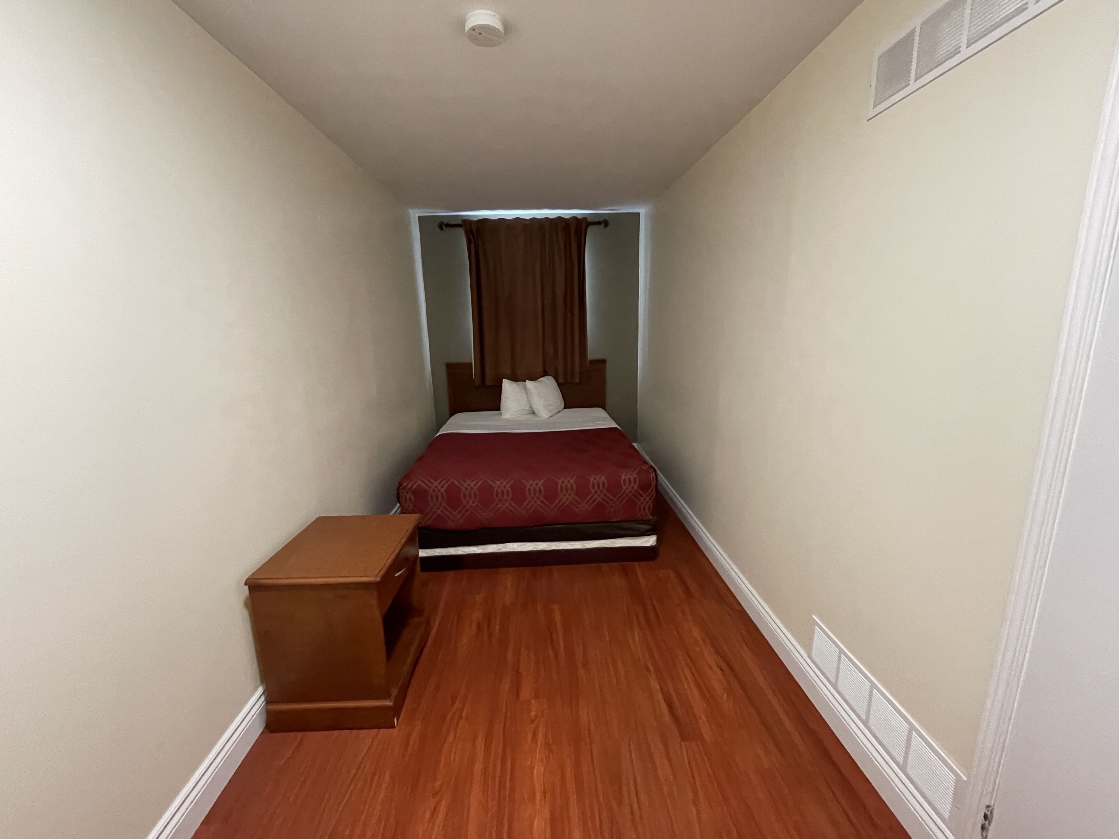 Una habitación de hotel estrecha con una cama, una mesita de noche y una pequeña mesa de madera.