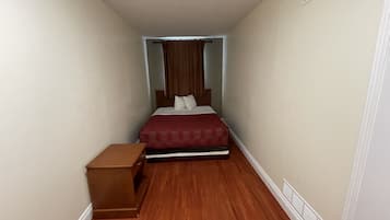 Una habitación de hotel estrecha con una cama, una mesita de noche y una pequeña mesa de madera.