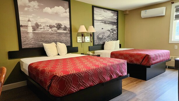 Habitación de hotel con dos camas, cada una con una colcha estampada roja y almohadas blancas. Hay dos lámparas de pared, un cuadro grande con una imagen de un faro y un equipo de aire acondicionado en la pared.