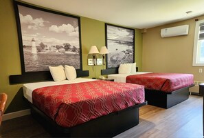 Habitación de hotel con dos camas, cada una con una colcha estampada roja y almohadas blancas. Hay dos lámparas de pared, un cuadro grande con una imagen de un faro y un equipo de aire acondicionado en la pared.