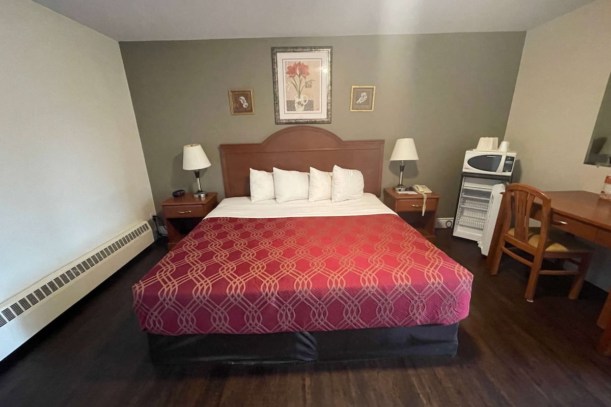 A hotel room with a large bed, two bedside tables with lamps, a microwave, a small refrigerator, a wooden chair, and a framed picture on the wall.