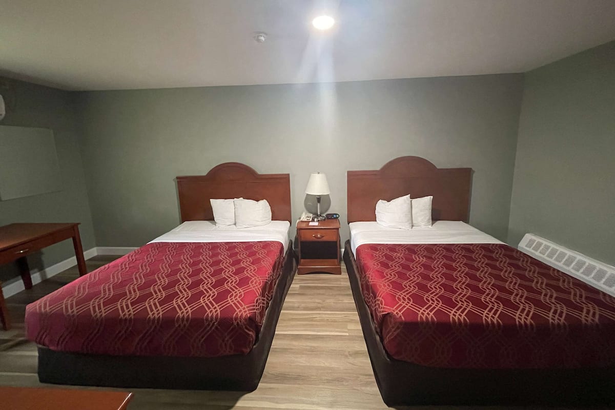 A hotel room with two beds, each with a wooden headboard and red patterned bedspreads. There is a nightstand between the beds with a lamp and a phone.
