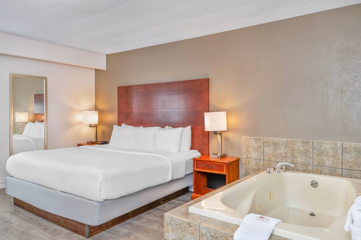 Een hotelkamer met een groot bed, een houten hoofdeinde, twee bedlampen, een nachtkastje, een badkamer met een bad en een betegelde douchewand.