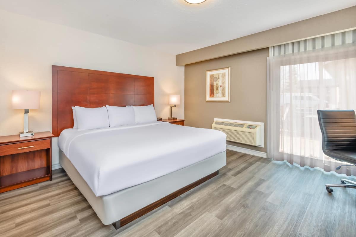 Een hotelkamer met een groot bed, nachtkastjes met lampen, een stoel, een radiator en een raam met dunne gordijnen.