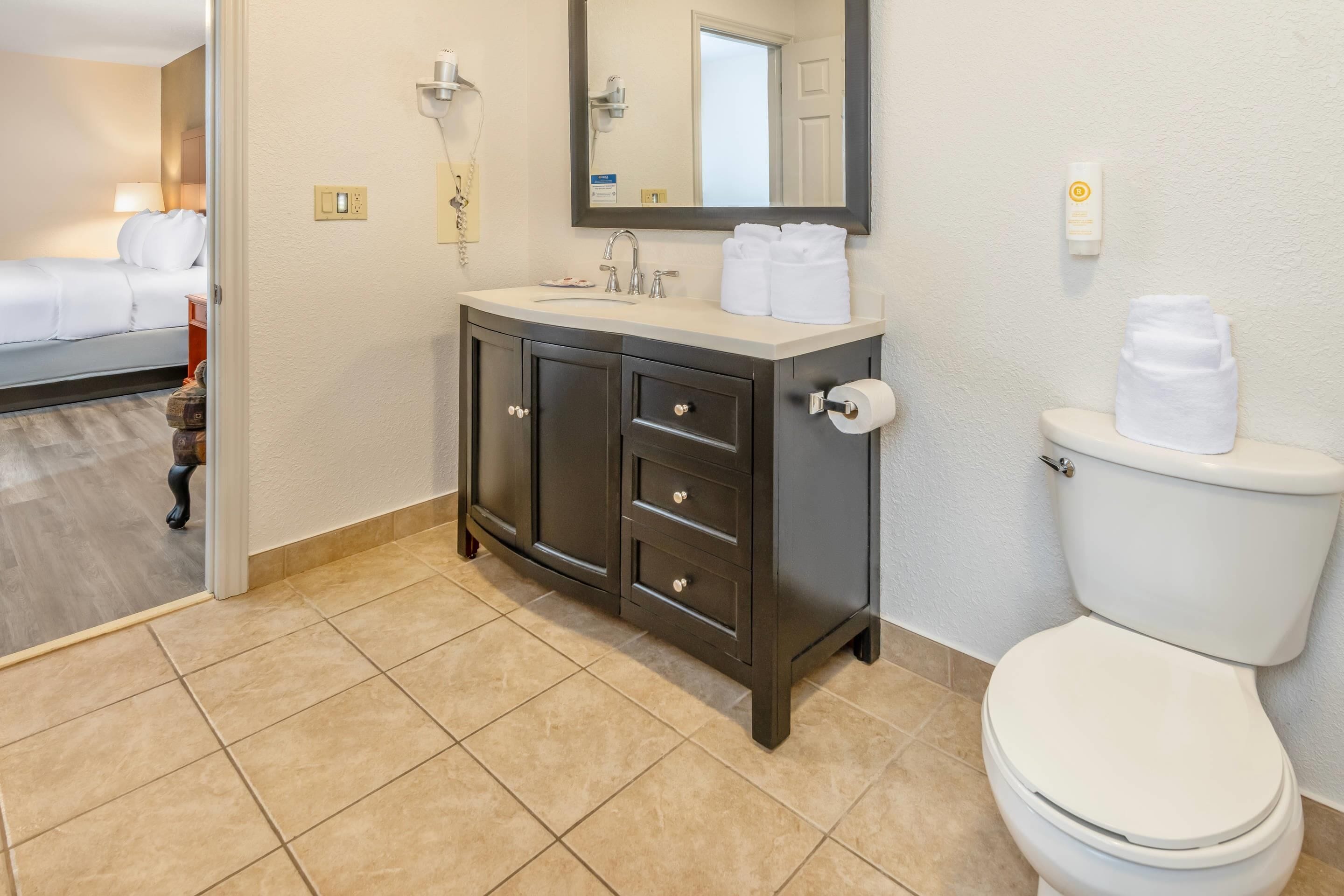 Un bagno d'hotel con un water bianco, un mobile da bagno in legno scuro con cassetti, uno specchio sopra il lavandino e un letto visibile nella stanza adiacente.